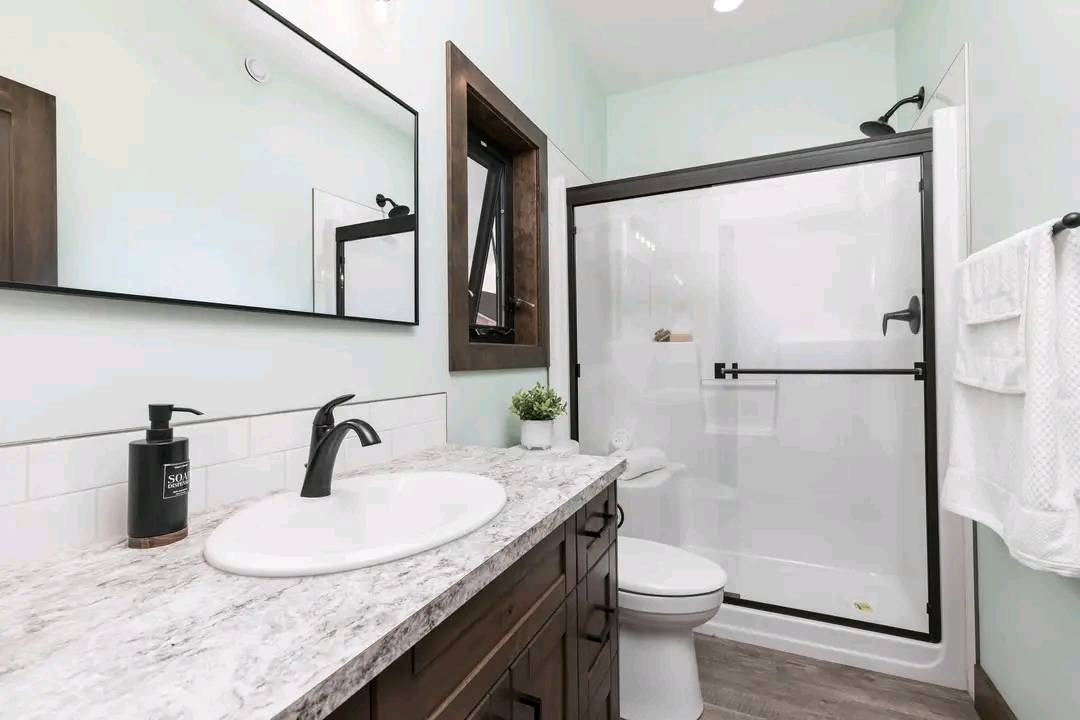 A modern bathroom with a glass-enclosed shower, marble countertop, dark wood cabinets, and a black faucet. A small plant and soap bottle sit on the counter.