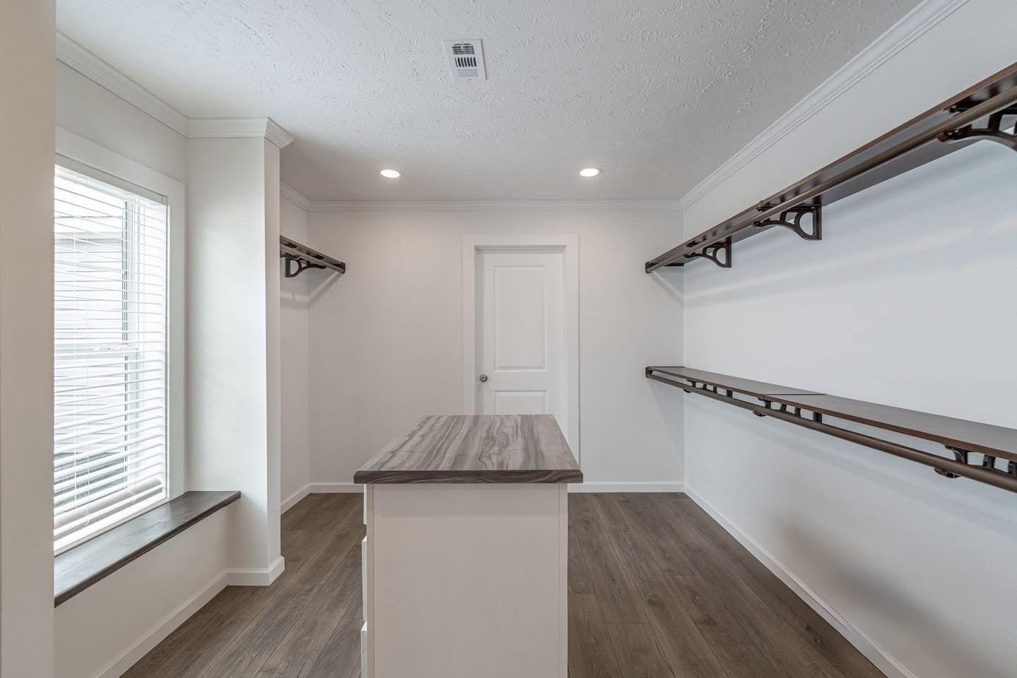 Bright, minimalist closet with white walls, wood flooring, and a central island. Long shelves line both sides, and natural light filters through a window.