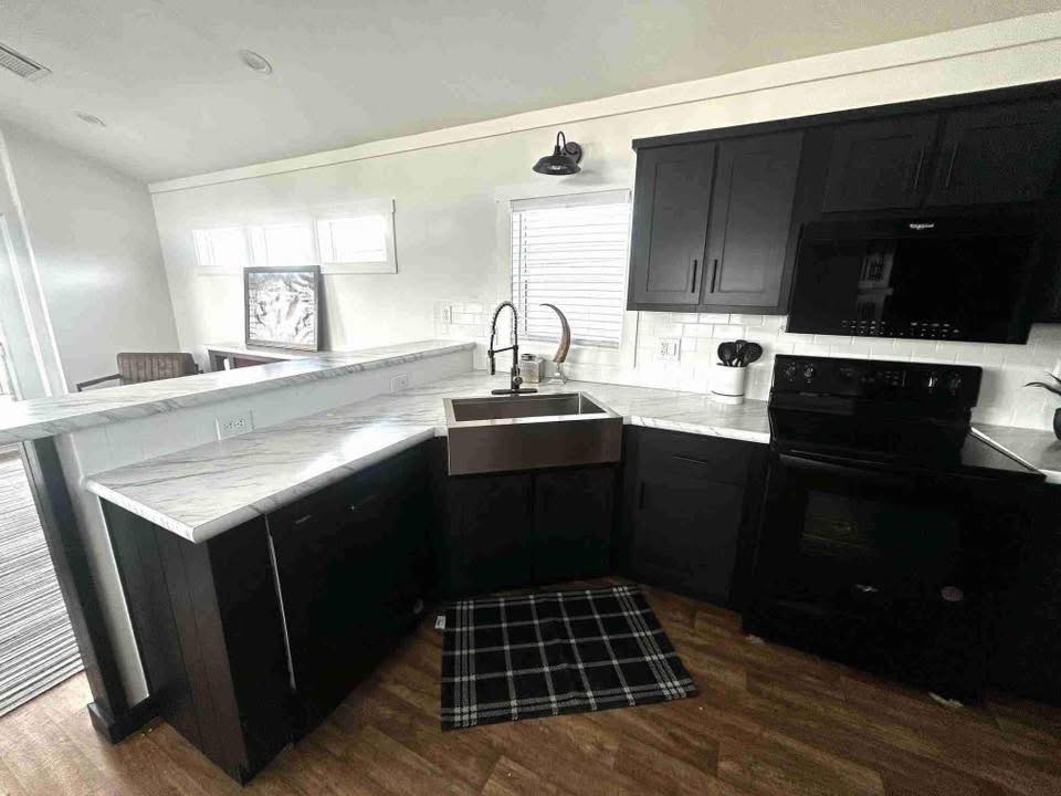 Modern kitchen with dark cabinets, marble countertops, and a farmhouse sink. A black stove and plaid rug add contrast. Bright, airy ambiance.