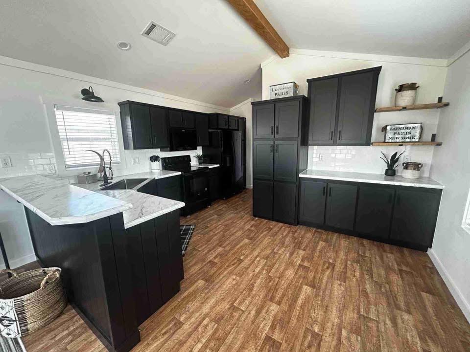 Modern kitchen with dark cabinets, white marble countertops, and wooden floors. High ceiling with a beam, minimal decor, and a cozy, elegant vibe.