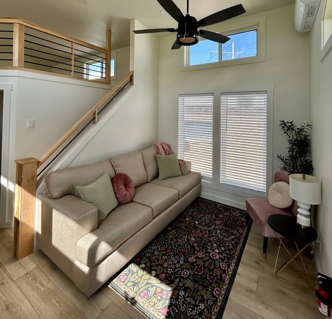 Modern living room with beige sofa, pastel cushions, floral rug, and a sleek black ceiling fan. Sunlit space with large windows and minimalist decor.