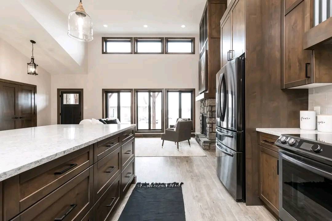 Modern kitchen with wooden cabinets, marble countertops, and stainless steel appliances. Large windows and high ceilings create an open, airy feel. Cozy seating area visible.