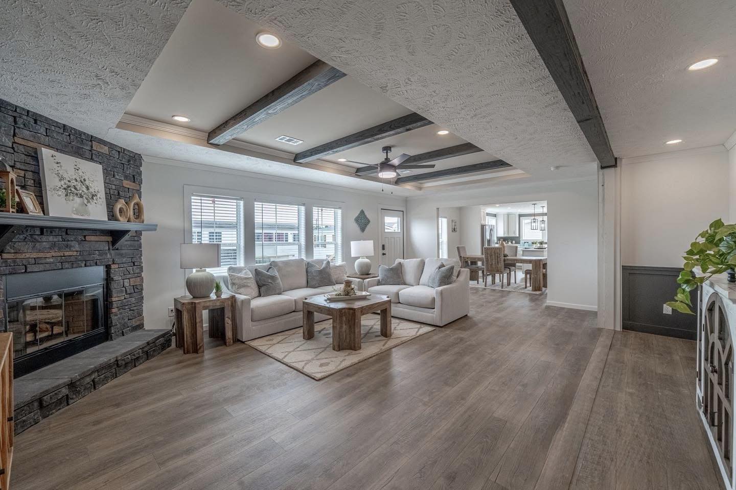Spacious living room with light wood floors, white sofas, and a large stone fireplace. Exposed wooden beams and soft lighting create a cozy ambiance.