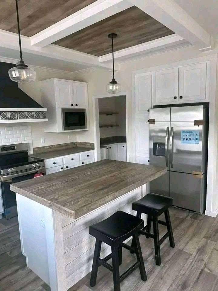 Modern kitchen with a wooden island, two black stools, stainless steel fridge, and white cabinets. Pendant lights add a cozy, inviting feel.
