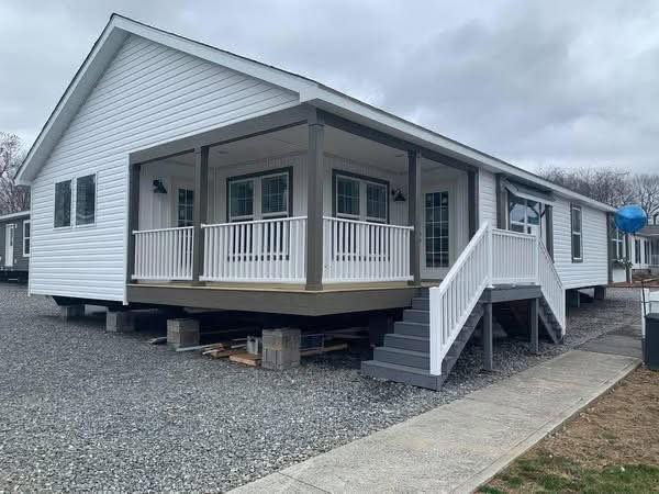 A white modular home with a front porch and steps, elevated on blocks. It sits on a gravel lot under a cloudy sky, conveying a sense of new construction.