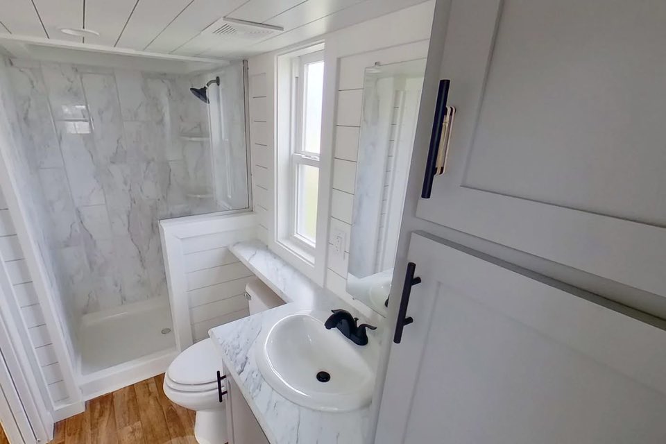 Small bathroom with wood floor features a marble-tiled shower, white sink with black faucet, and cabinet. Bright light from window creates airy feel.