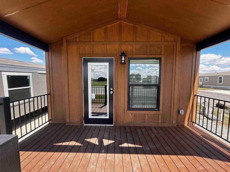 A wooden porch features a door with glass panels and a window, both framed in black. Iron railings and a light fixture add contrast under a blue sky.
