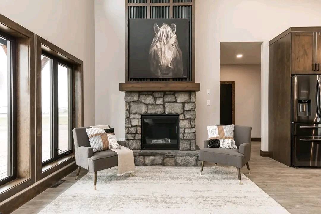 Elegant living room with a stone fireplace, horse portrait above. Two gray chairs with cushions and throws flank the fireplace. Large windows and wood accents add warmth.