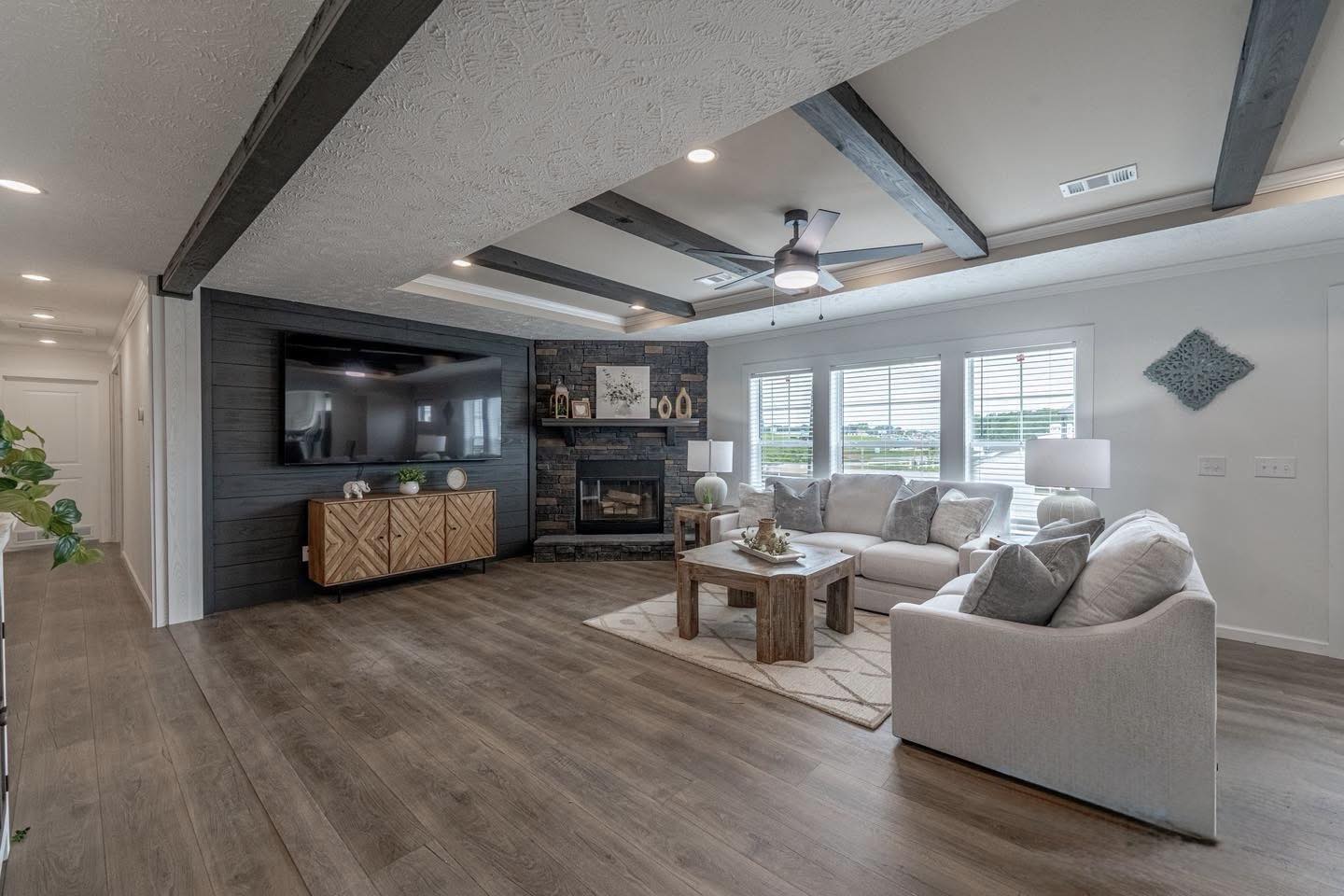 Modern living room with wood beams, neutral tones, and large windows. Features a sectional sofa, wooden table, fireplace, and wall-mounted TV. Cozy ambiance.