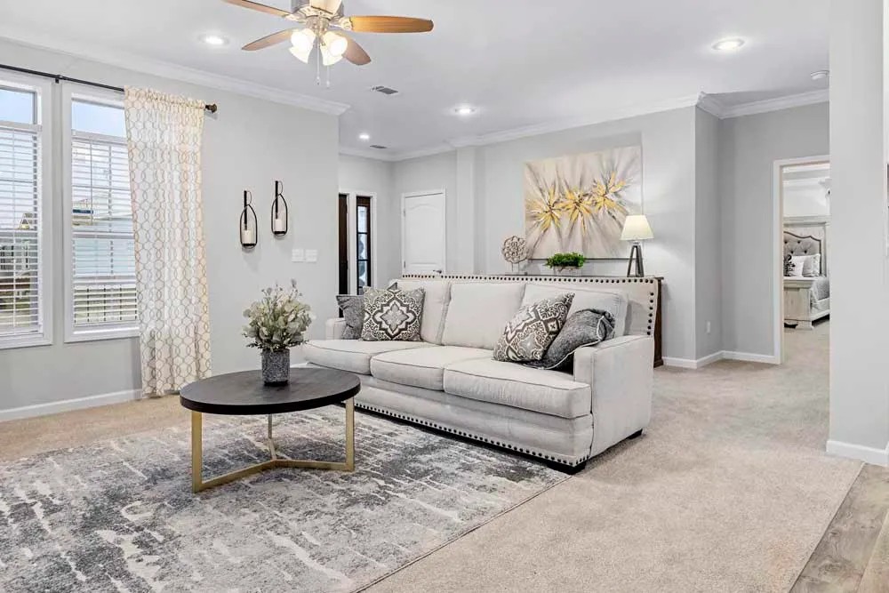 Modern living room with light gray walls, a cream sofa with patterned cushions, round coffee table, abstract art, and a ceiling fan. Bright and inviting ambiance.