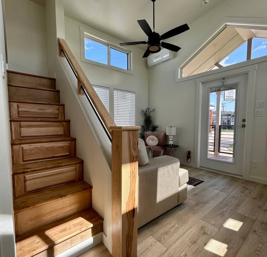 A cozy, sunlit living area features a wooden staircase, beige sofa, and modern black ceiling fan. Large windows and glass door enhance the airy feel.