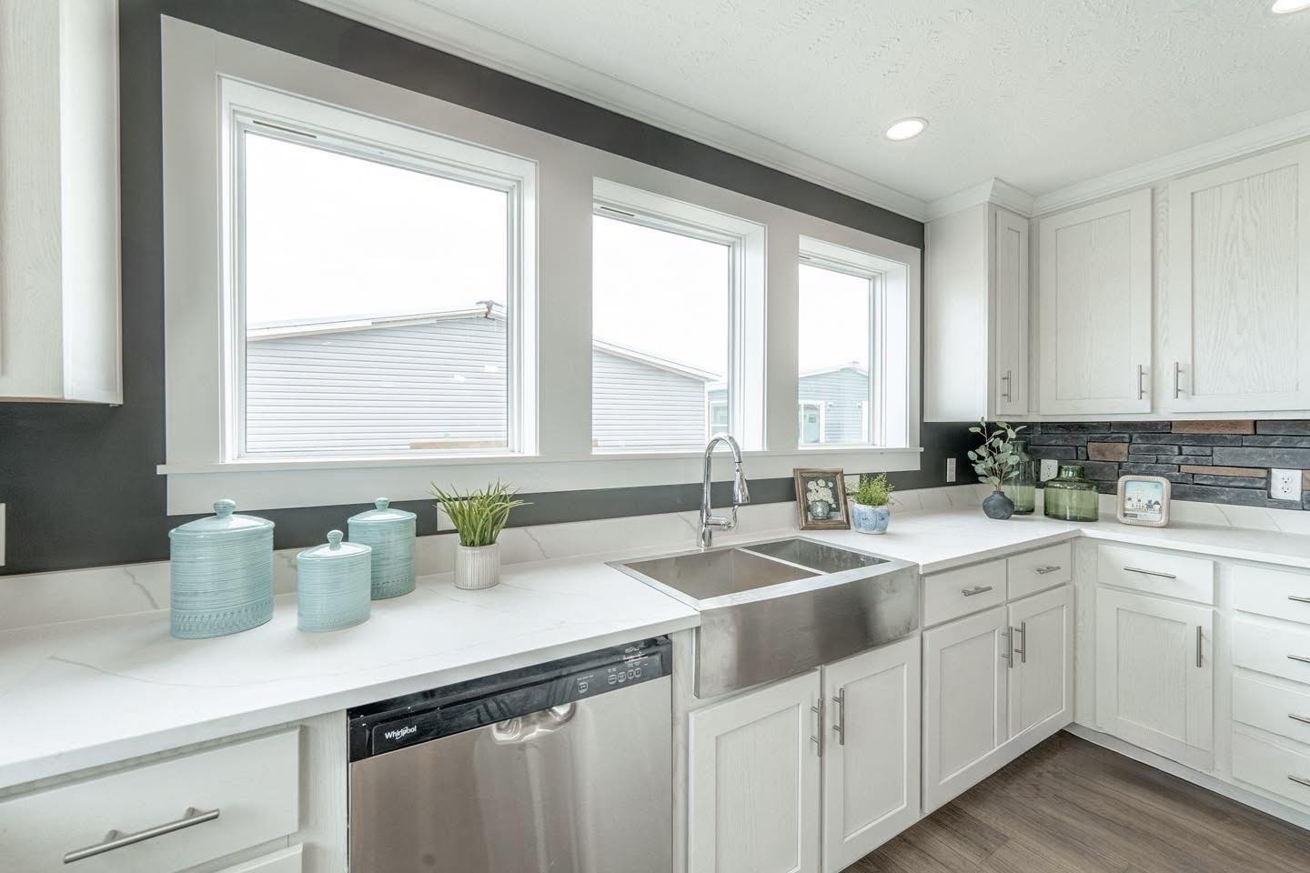 Bright kitchen with white cabinets, a stainless steel sink, dishwasher, and large windows. Decor includes light blue jars and plants on the counter.