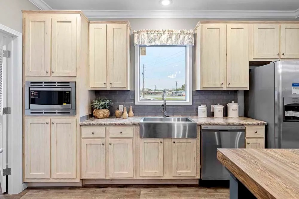 A cozy kitchen features light wooden cabinets, a stainless steel farmhouse sink, and modern appliances. A large window provides natural light.