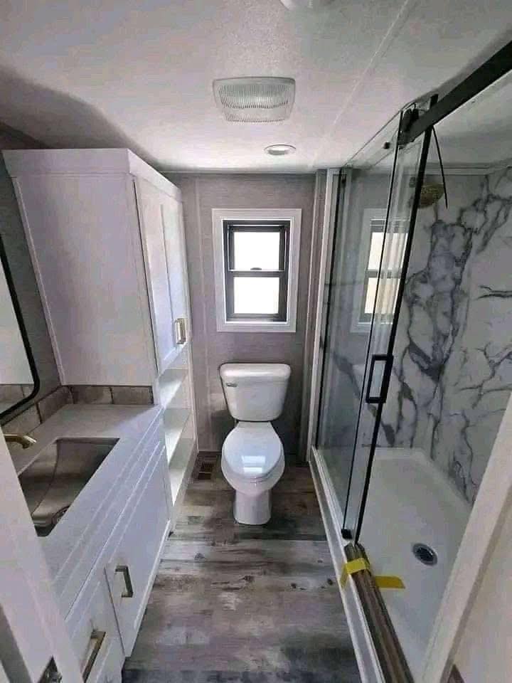 Narrow bathroom with toilet, small window, sleek white vanity on the left, and a shower with glass doors and marble-patterned walls on the right.