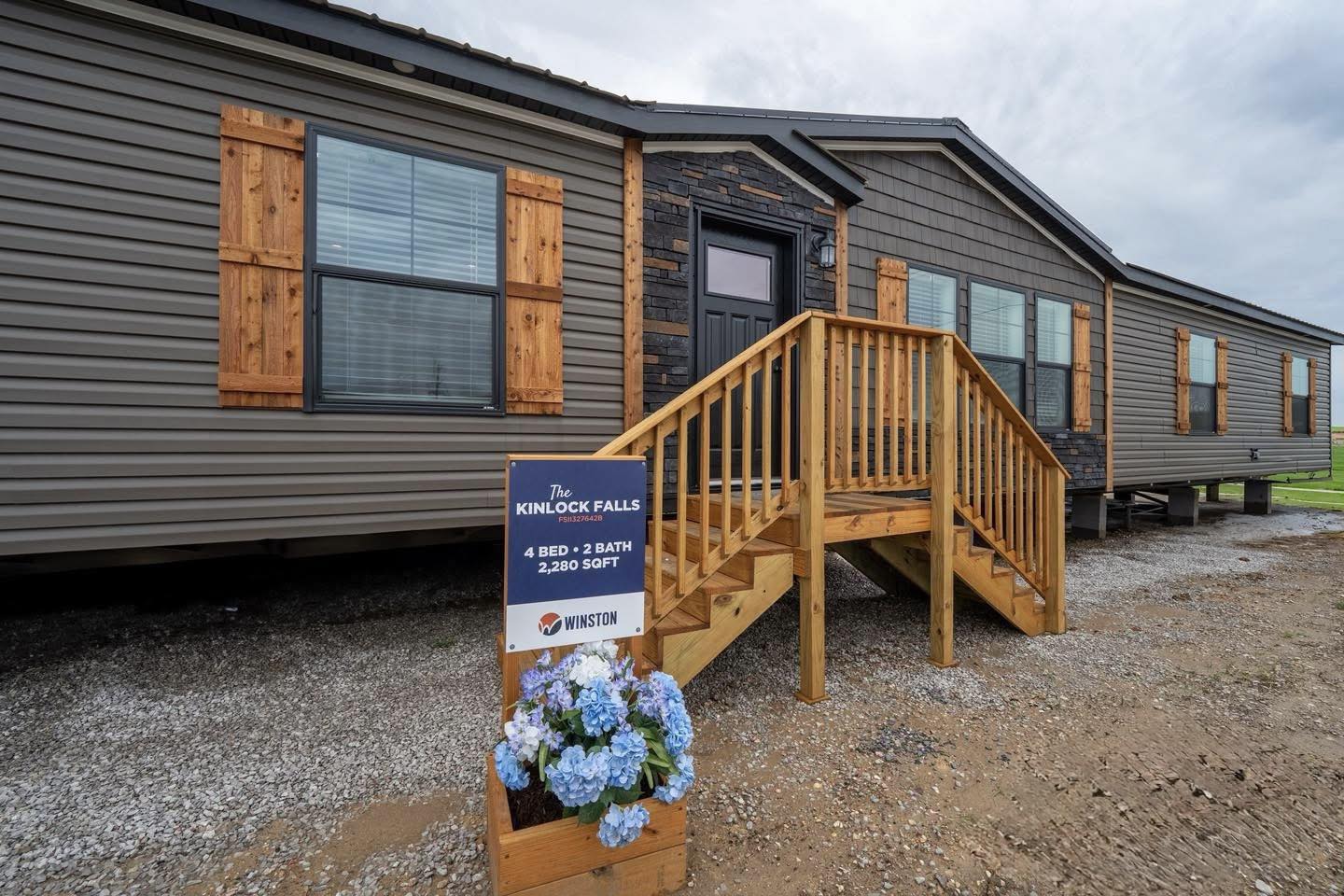 A modern manufactured home with gray siding and wooden shutters, featuring a small wooden staircase. A sign reads "Kinlock Falls, 4 bed, 2 bath, 2,280 sqft." Blue flowers in a planter create a welcoming feel.