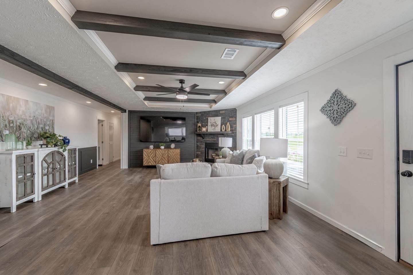 Spacious living room with a modern design features a beige sofa, wooden floor, ceiling beams, large TV, and windows with abundant natural light.