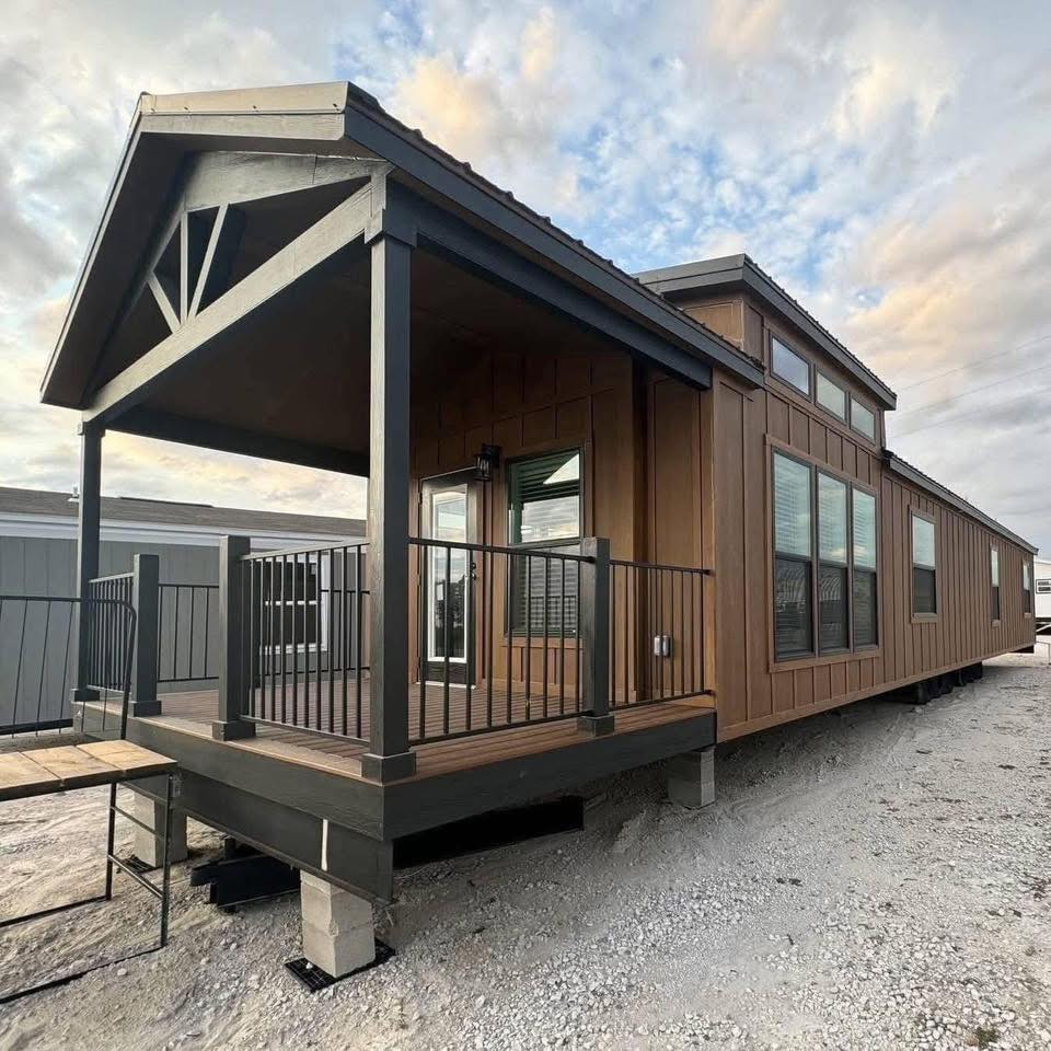 A modern brown cabin with a gabled roof sits on raised foundations. It features large windows and a black railing porch under a cloudy sky.