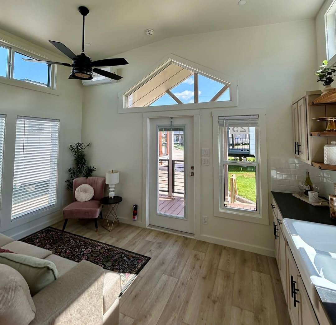 Bright living area with light wood floors, a beige sofa, and a pink chair by a tall plant. Large windows and glass door lead to a sunny patio. Ceiling fan above, creating an airy feel.