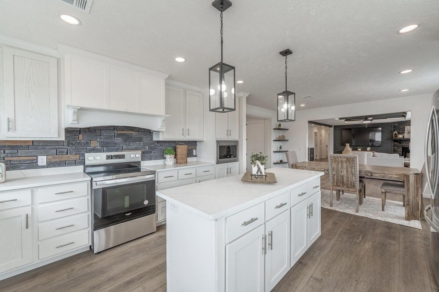 Spacious modern kitchen with white cabinets, stainless steel appliances, black tile backsplash, and pendant lights. Wooden dining area visible in background.