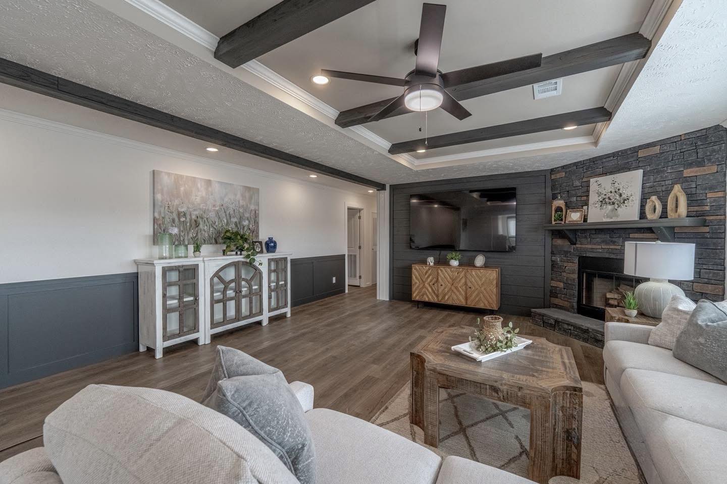 Modern living room with neutral tones, featuring a large ceiling fan, light sofas, a wooden coffee table, and a stone fireplace. Warm, inviting atmosphere.