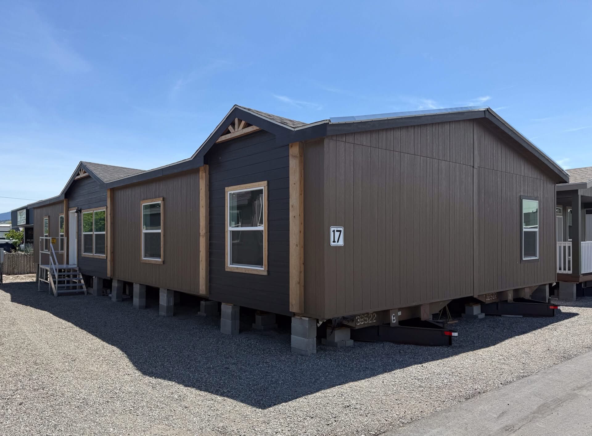 A large modular home with brown siding sits on a gravel lot under a clear blue sky. Number 17 is displayed on one side, emphasizing its placement.