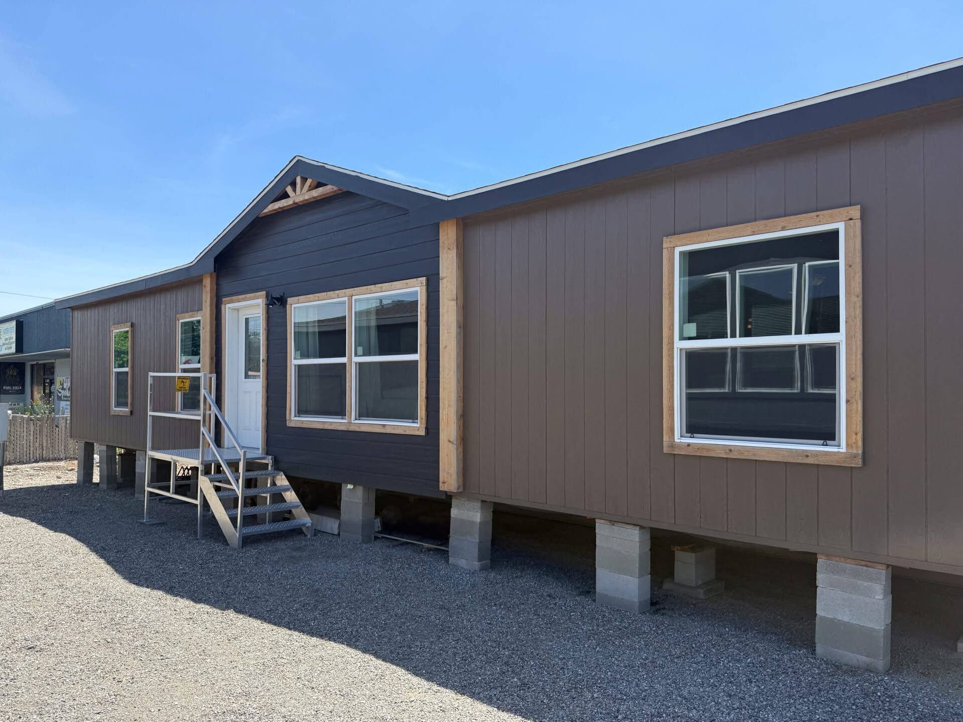 A brown and dark gray modular home elevated on cinder blocks with white framed windows and a small porch. The sky is clear and sunny.