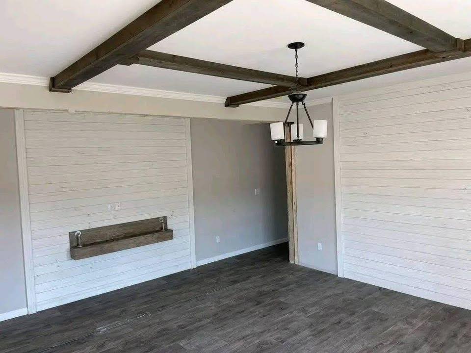 Cozy room with wood panel walls, wooden ceiling beams, and a modern chandelier. The floor is dark wood, creating a warm, rustic ambiance.