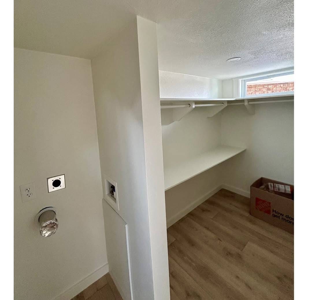 Small, empty laundry room with light wood flooring, white walls, and a shelf with hanging rods. A cardboard box sits on the floor in the corner.