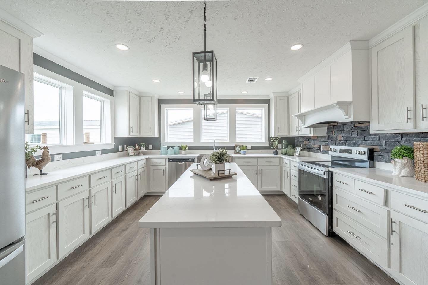 Bright, modern kitchen with white cabinetry and countertops, dark backsplash, and stainless steel appliances. Central island with plants, wood floor. Elegant and serene atmosphere.