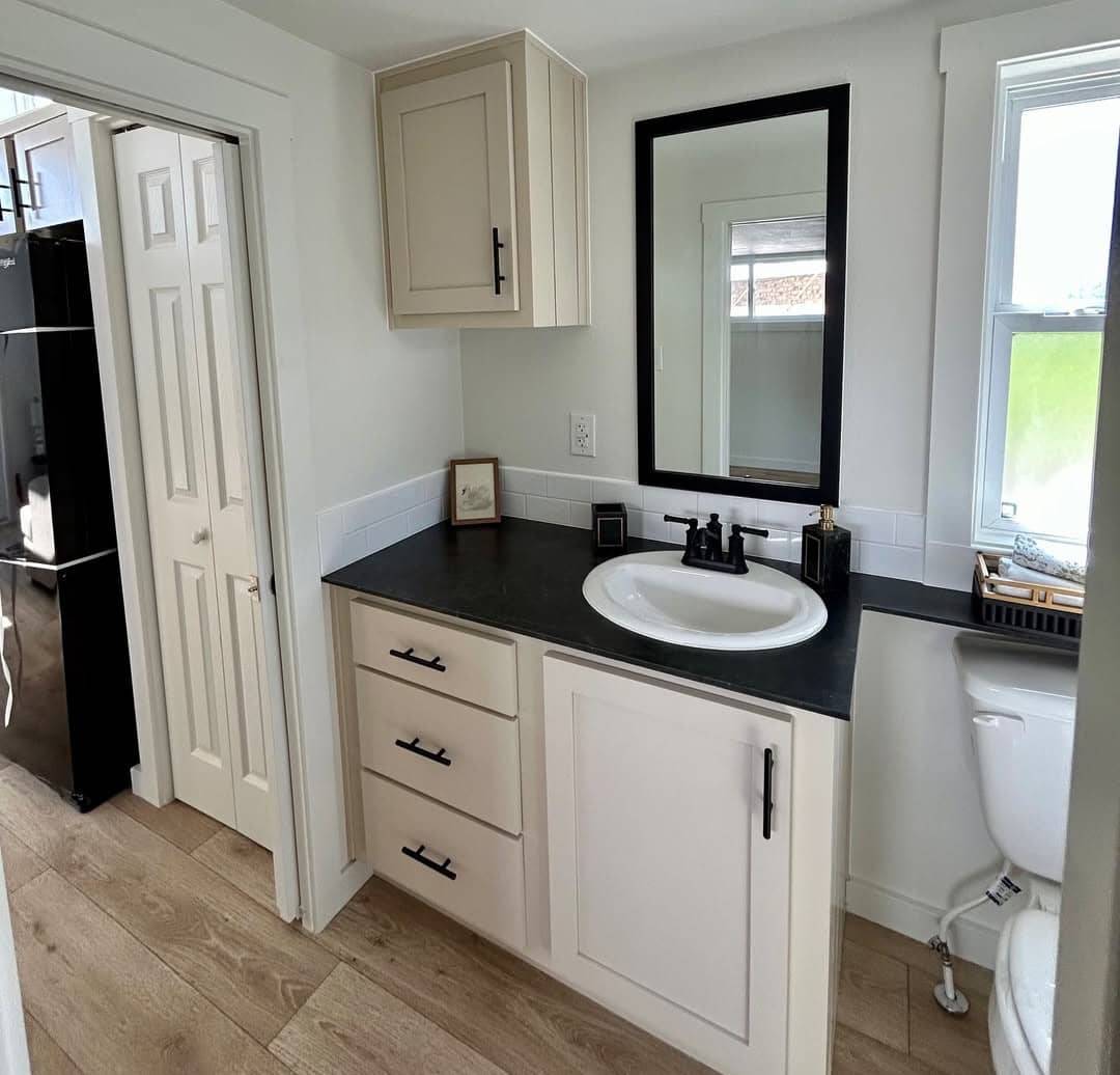 A small, modern bathroom with light wood flooring and beige cabinetry. Features a black-countertop vanity, black faucet, mirrored wall, and white accents, creating a clean, minimalist feel.