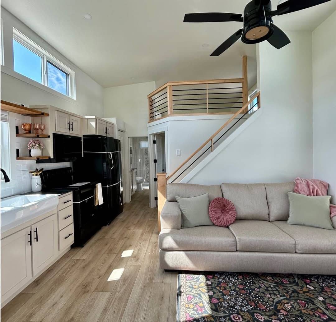 Stylish tiny home interior with loft. Features a beige sofa, decorative pillows, and a modern kitchen with black appliances. Bright and cozy ambiance.