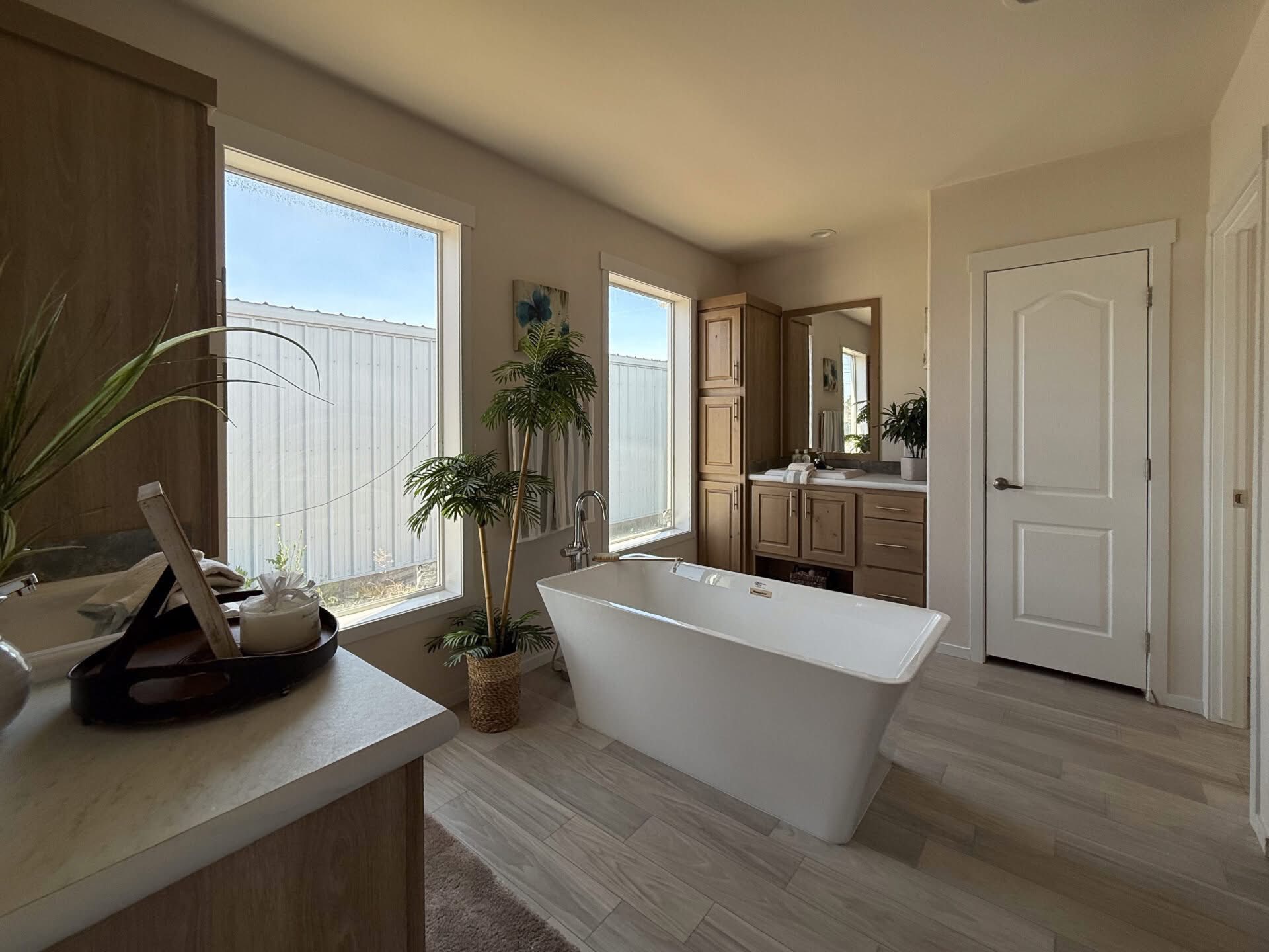 Bright modern bathroom with a white freestanding tub, large windows, wooden cabinets, and indoor plants. The atmosphere is calm and inviting.