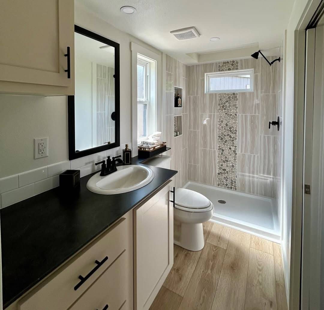 Modern bathroom with wood flooring, a black countertop, rectangular sink, and a large mirror. Shower features stone accent tiles and ample natural light.
