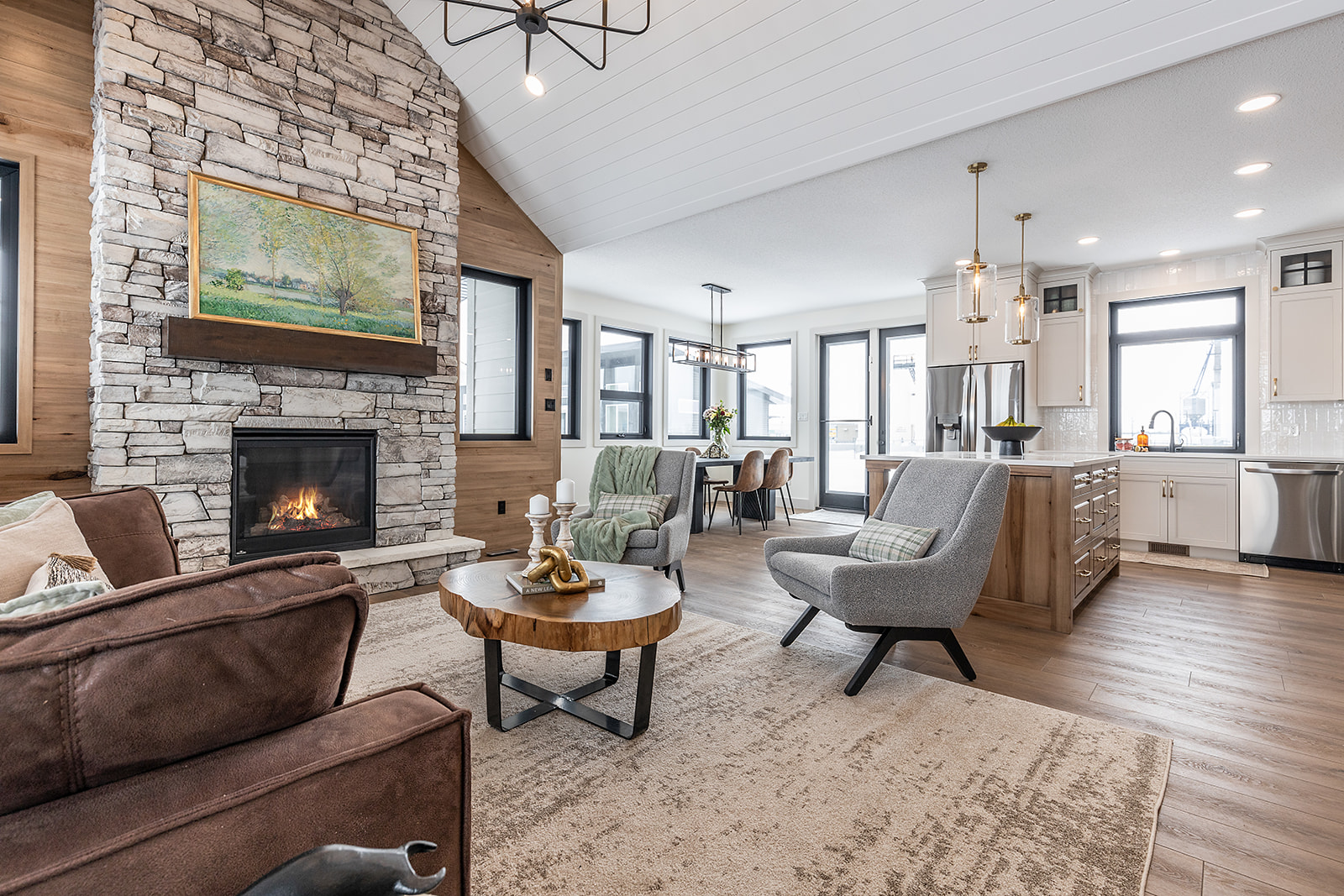 Spacious living room with stone fireplace, modern furniture, and cozy décor. Open layout leads to a bright kitchen and dining area with stylish lighting.