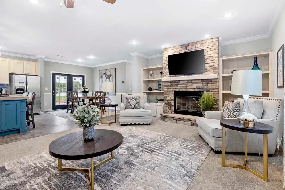 Spacious living room with gray sofas, a stone fireplace, and a mounted TV. Light decor, round coffee tables, and an elegant dining area create a cozy ambiance.