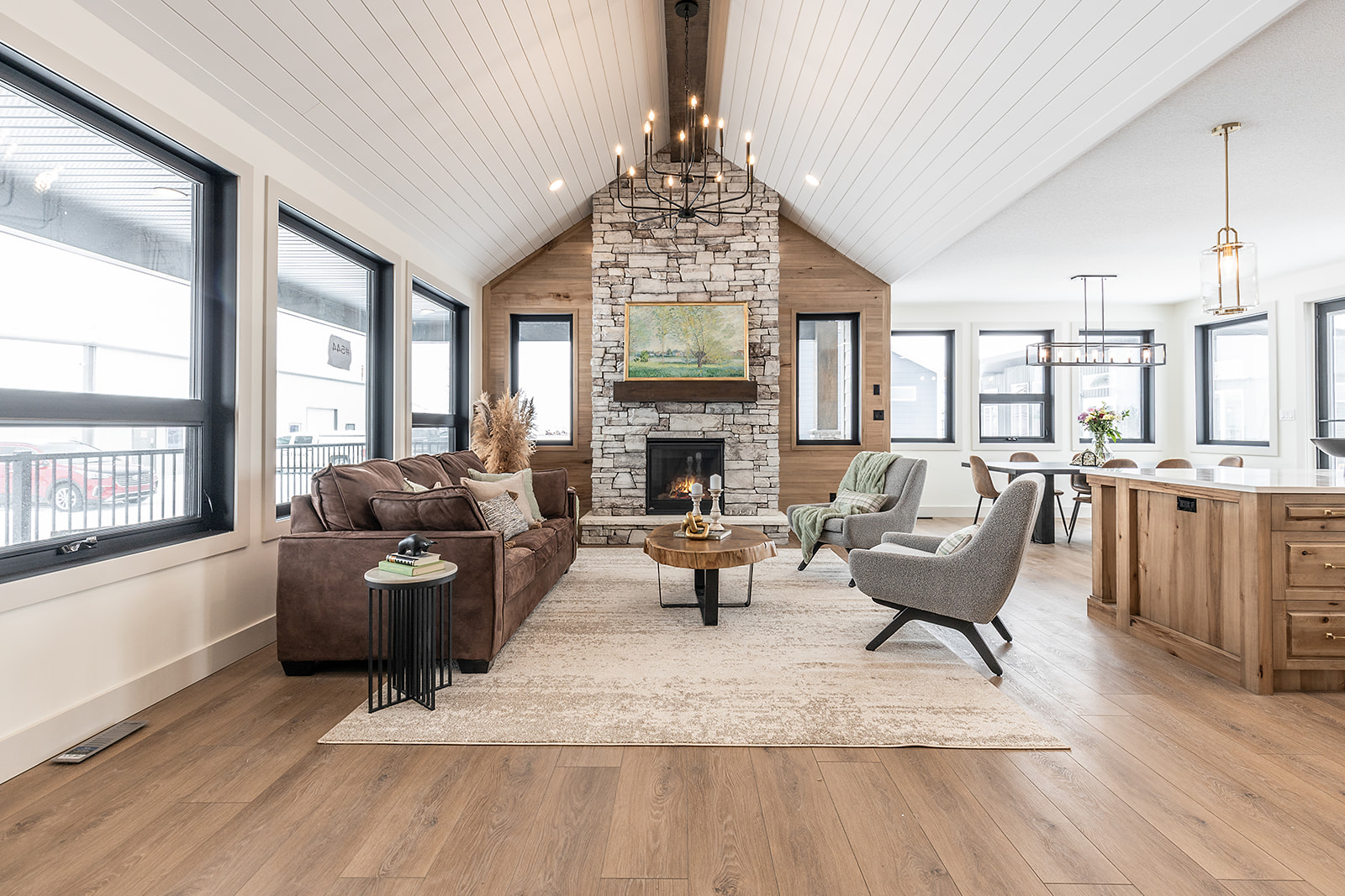 Spacious living room with vaulted ceiling and chandelier. Features include a stone fireplace, brown sofa, gray chairs, wood accents, and rustic decor. Cozy and elegant.
