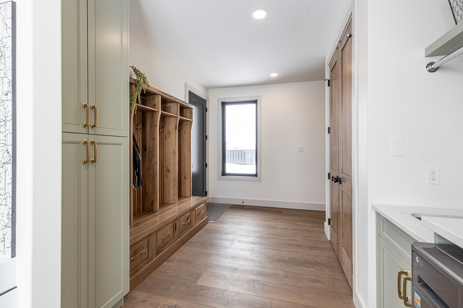 Mudroom with wooden floors, light green cabinets, and a wooden bench with coat hooks and cubbies. A large window and natural light create an airy feel.