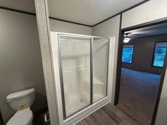 Bathroom with a glass-enclosed shower, beside a toilet on a wooden floor. An adjoining room is visible, featuring carpet and a ceiling fan.