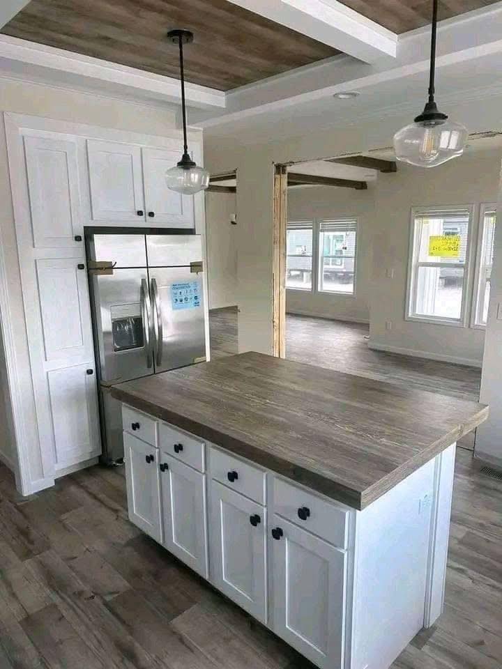Modern kitchen with white cabinets and large island featuring a wood countertop. Stainless steel fridge, pendant lights, and wood floors add warmth.