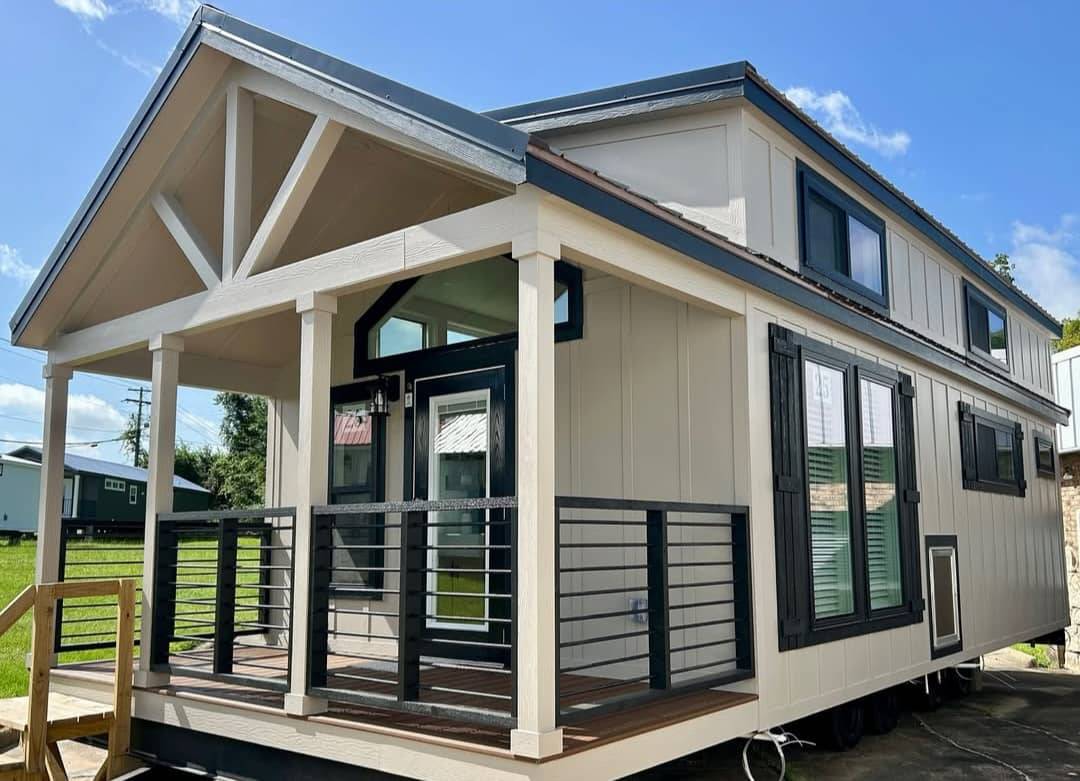 Modern tiny house with a beige exterior and dark trim, featuring a small covered porch with railings. The clear blue sky adds a cheerful tone.