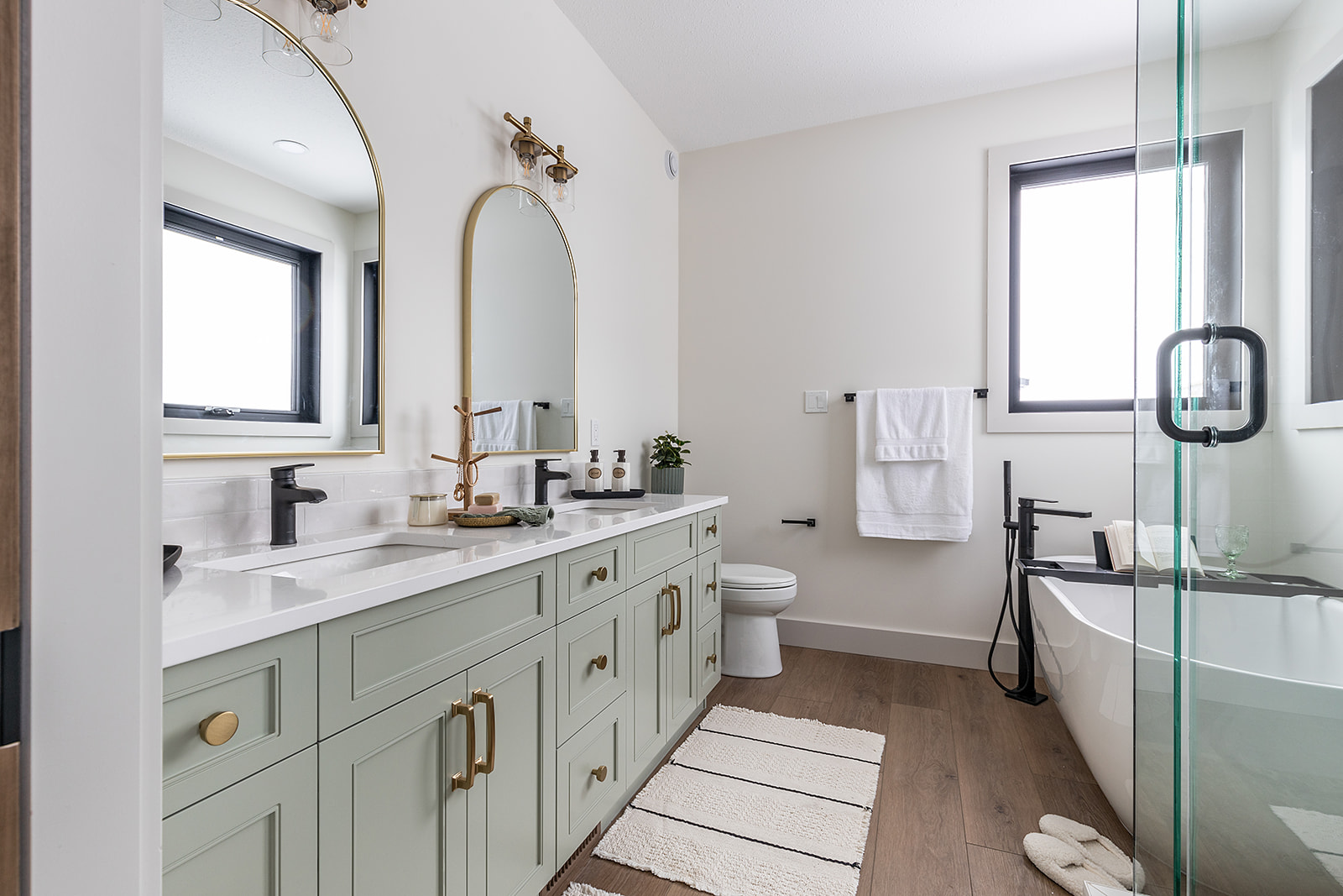 Modern bathroom with green vanity features dual sinks, large mirrors, gold accents, and elegant fixtures. A freestanding tub sits by large windows.