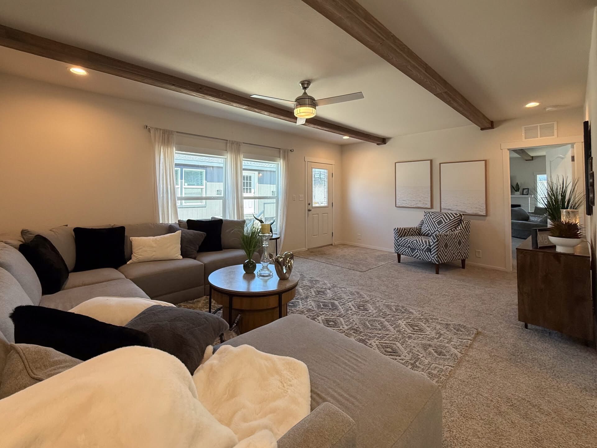 Spacious living room with gray sectional sofa, patterned armchair, and round wooden table. Neutral tones and natural light create a cozy, welcoming feel.
