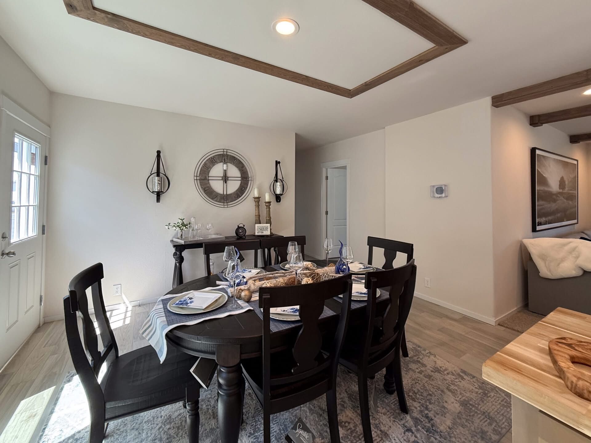 Dining room with a rustic wooden table set for six, elegant tableware, and a centerpiece. Decor includes a round wall clock and candles, evoking a cozy atmosphere.