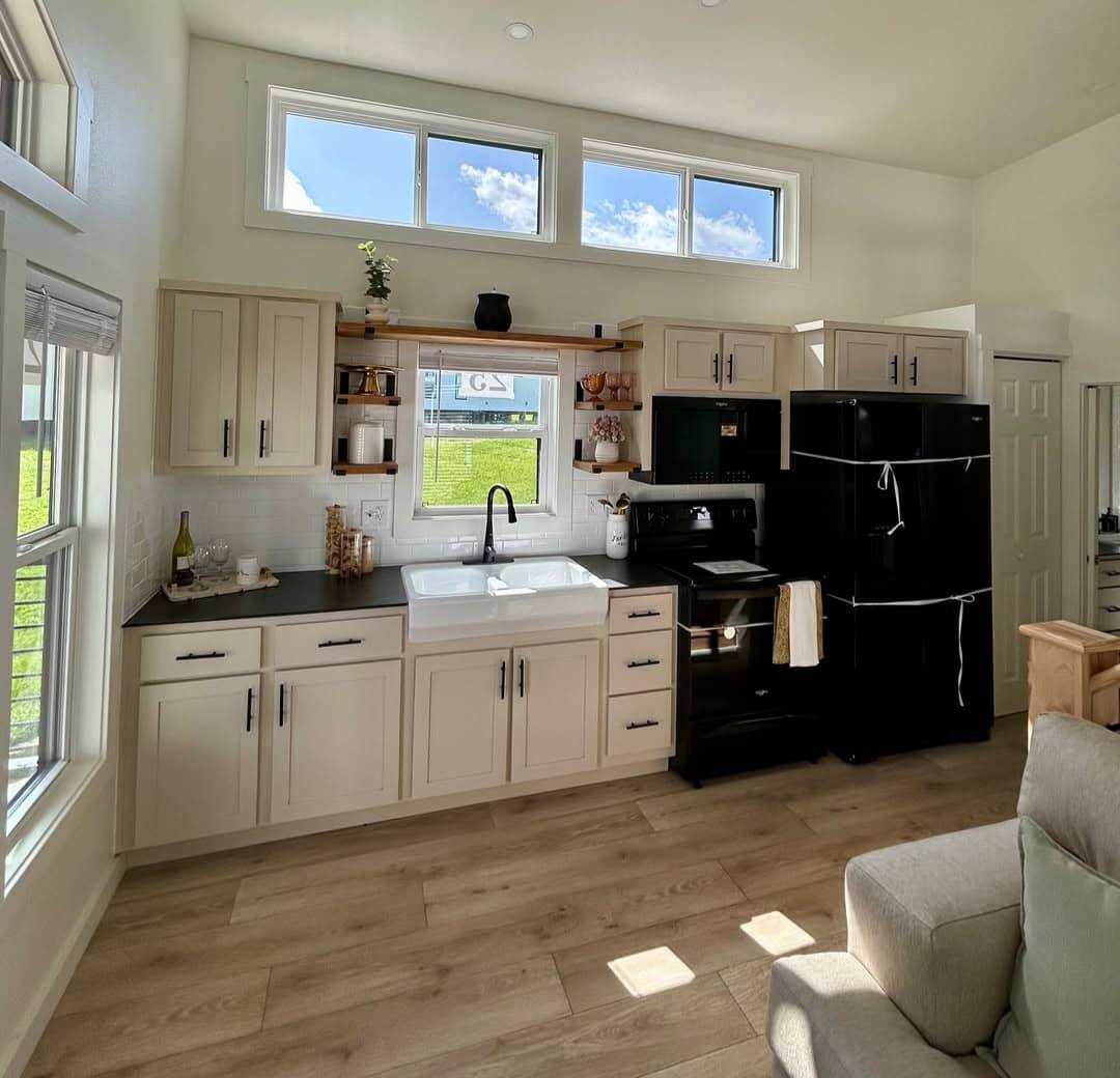 Bright, airy kitchen with beige cabinets, black countertops, and light wood floors. Sunlight streams through tall windows, creating a cozy, welcoming vibe.