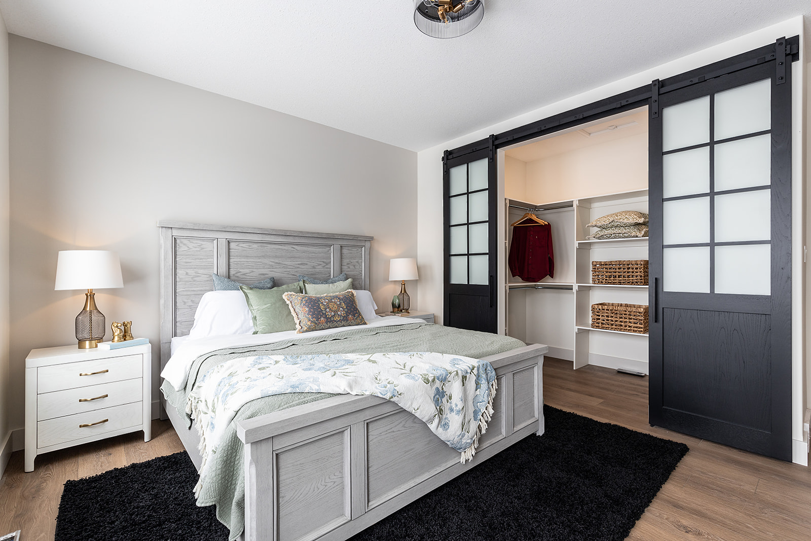 A cozy bedroom with a light gray wooden bed, floral bedding, and two lamps on white nightstands. Open closet with sliding black doors and wicker baskets.