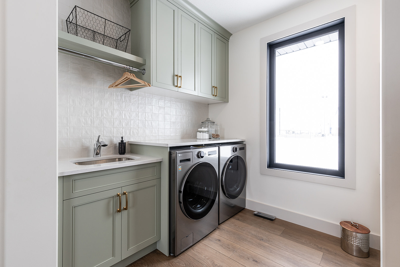 Modern laundry room with light green cabinets, stainless steel washer and dryer, a small sink, and a large window. Clean, bright, and organized space.