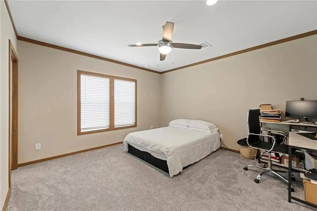 Bedroom with beige walls and carpet, featuring a neatly made bed. A desk with a computer, chair, and papers is in the corner. Ceiling fan overhead.