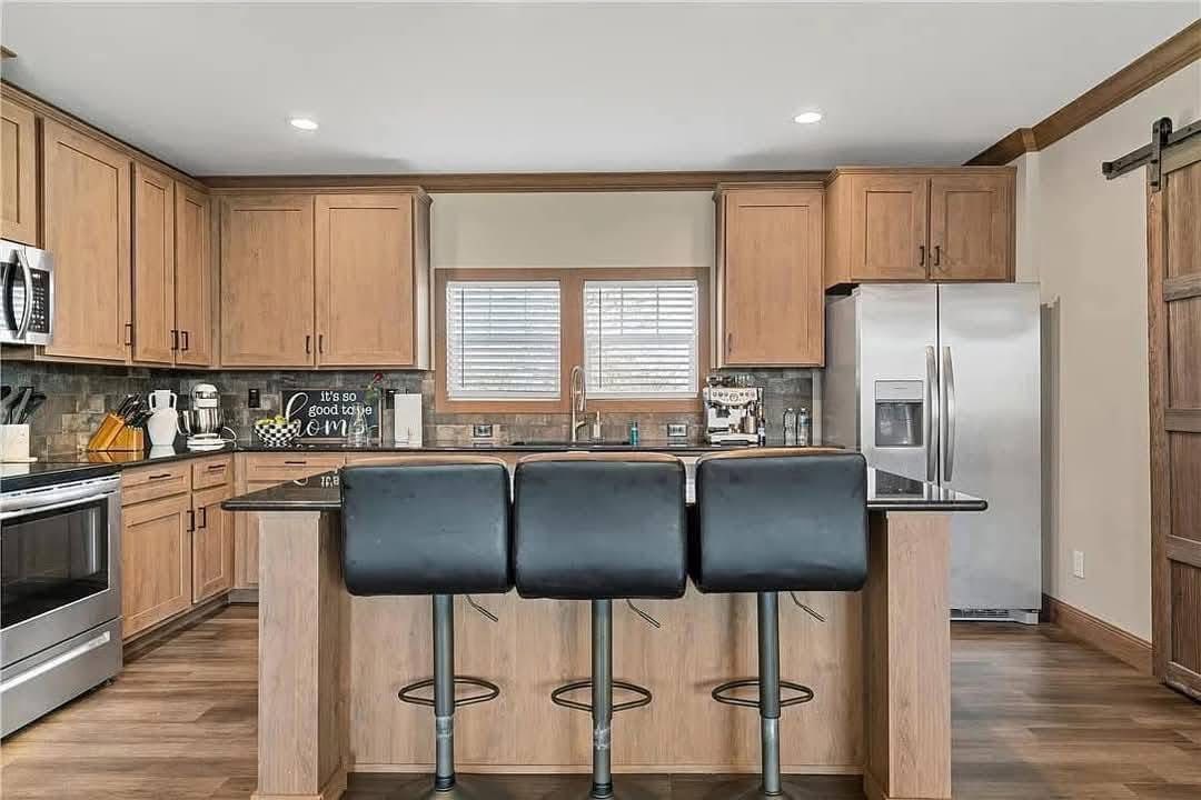 Modern kitchen with wooden cabinets, stainless steel appliances, and three black bar stools at a central island. Warm, cozy atmosphere with ample lighting.