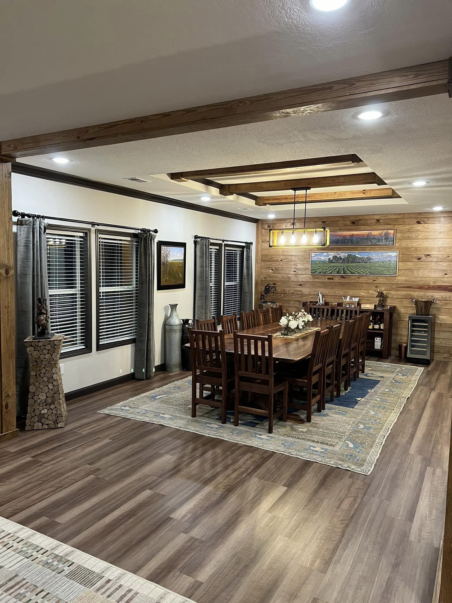 Warm, rustic dining room with a long wooden table set for twelve on a blue-patterned rug. Features wooden accents, soft lighting, and decorative art.