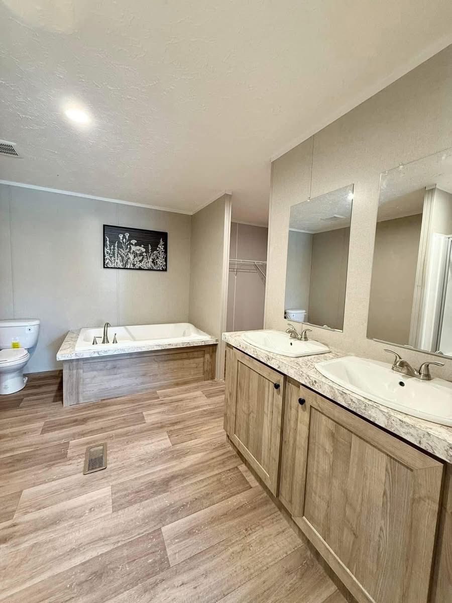 Spacious, modern bathroom with a dual-sink vanity and wood accents, a bathtub, toilet, and floral artwork. Light wood flooring adds warmth.